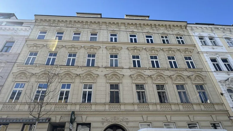 Historische Fassade eines Mehrfamilienhauses mit vielen Fenstern und Stuckverzierungen unter blauem Himmel.