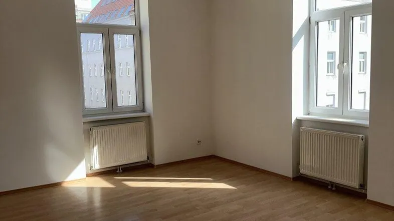 Helles, leeres Zimmer mit Parkettboden und großen Fenstern, das viel Tageslicht hereinlässt.