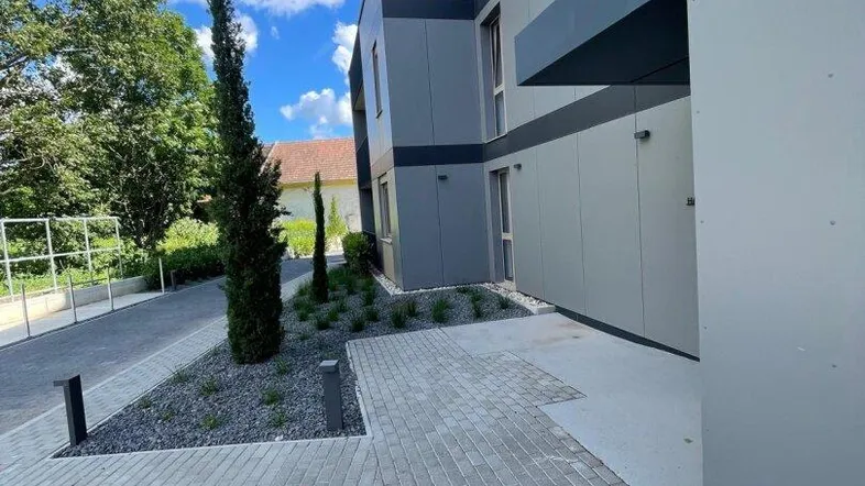 Moderne Gebäudefassade mit grauen Paneelen, gepflasterten Wegen und junger Bepflanzung unter blauem Himmel.