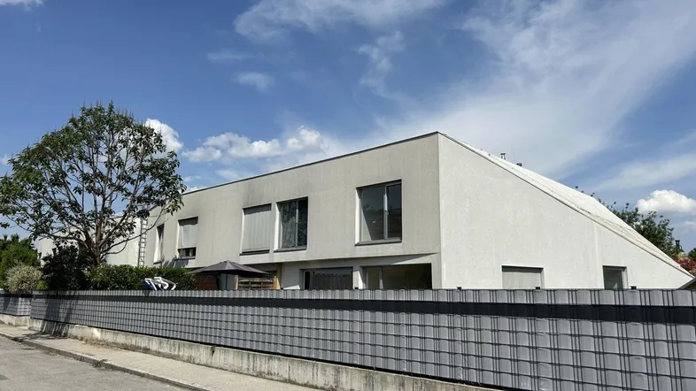 Moderne Wohnhausfassade mit großen Fenstern und einem Zaun im Vordergrund unter blauem Himmel.