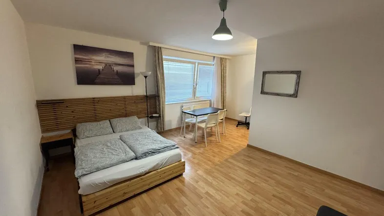 Helles Schlafzimmer mit Holzbett, kleinem Esstisch und Arbeitsbereich, ausgestattet mit Parkettboden.