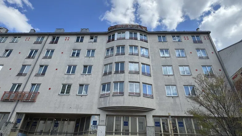 Fassade eines mehrstöckigen Wohn- und Geschäftshauses mit vielen Fenstern und kleinen Balkonen unter bewölktem Himmel.