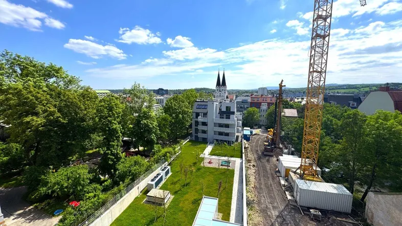 Blick auf die Baustelle, das neue Gebäude und die grüne Umgebung mit Kirchturm im Hintergrund.