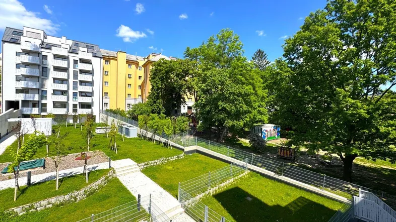 Gepflegte Gartenanlage mit jungen Bäumen und Wegen vor einem modernen Wohngebäude unter blauem Himmel.