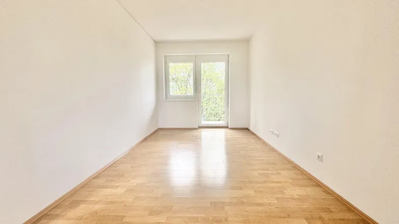 Helles, leeres Zimmer mit Parkettboden und Zugang zu einem Balkon.