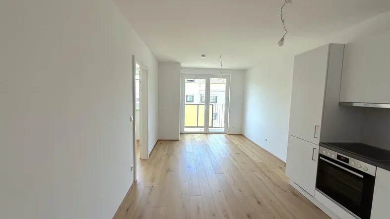 Helles Wohnzimmer mit integrierter Küchenzeile, Parkettboden und Zugang zum Balkon.