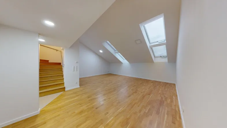 Heller Dachgeschossraum mit Holzfußboden, schrägen Wänden und mehreren Dachfenstern, daneben eine Holztreppe.