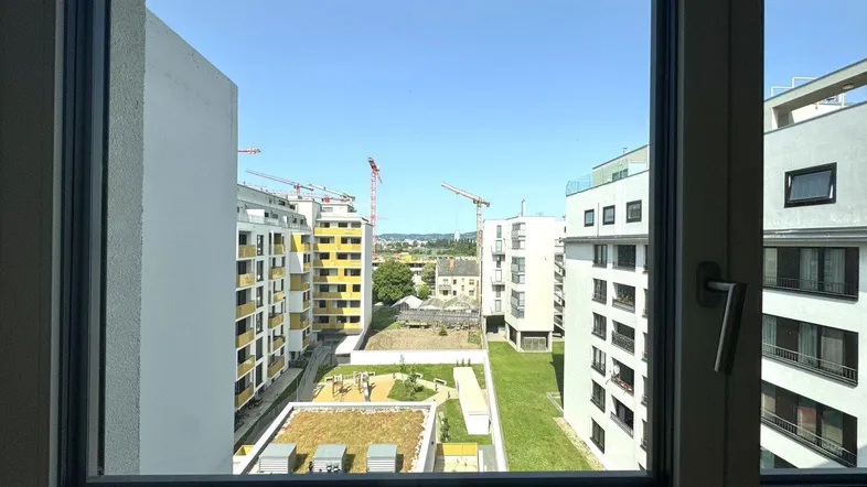 Blick aus dem Fenster auf moderne Wohngebäude, einen begrünten Innenhof mit Spielplatz und Baukräne.