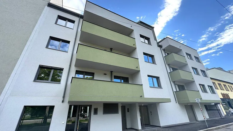 Moderne Fassade eines Neubaus mit hellen Wänden und grünen Balkonen unter blauem Himmel.