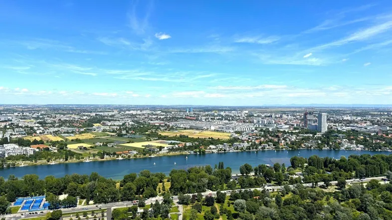 Weitläufige Luftaufnahme einer Stadtlandschaft mit Fluss, Grünflächen und urbaner Bebauung unter blauem Himmel.