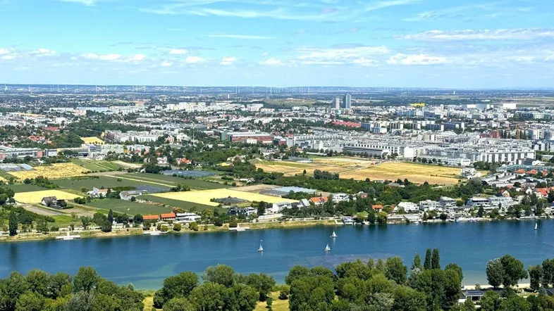 Luftaufnahme einer ländlichen Landschaft mit Fluss, Feldern, vereinzelten Häusern und Windkraftanlagen am Horizont.