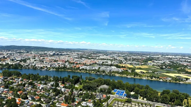 Weitläufige Luftaufnahme einer Stadtlandschaft mit Fluss, Wohngebieten und grünen Parkanlagen unter blauem Himmel.