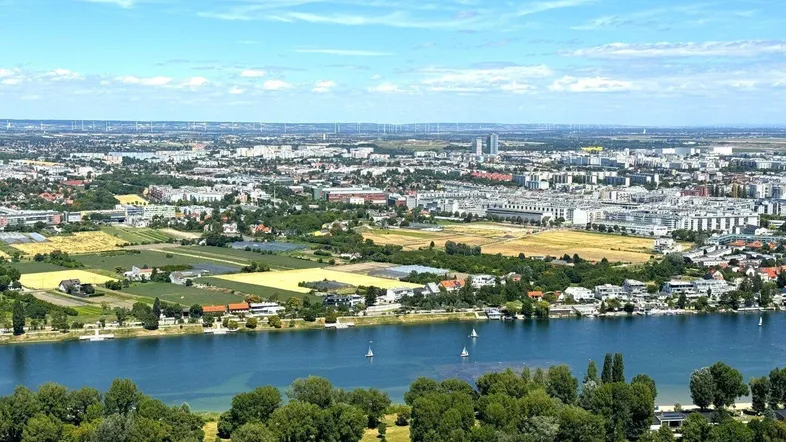 Weitläufige Luftaufnahme einer Stadtlandschaft mit Fluss, grünen Ufern und urbaner Bebauung unter blauem Himmel.