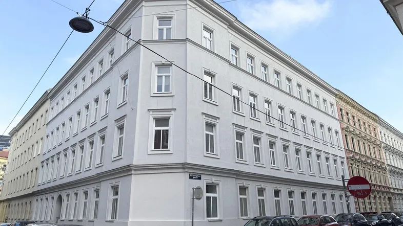 Graues Eckgebäude mit vielen Fenstern und parkenden Autos auf der Straße unter bewölktem Himmel.