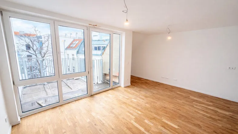Helles, leeres Wohnzimmer mit Parkettboden und Zugang zu einem Balkon.