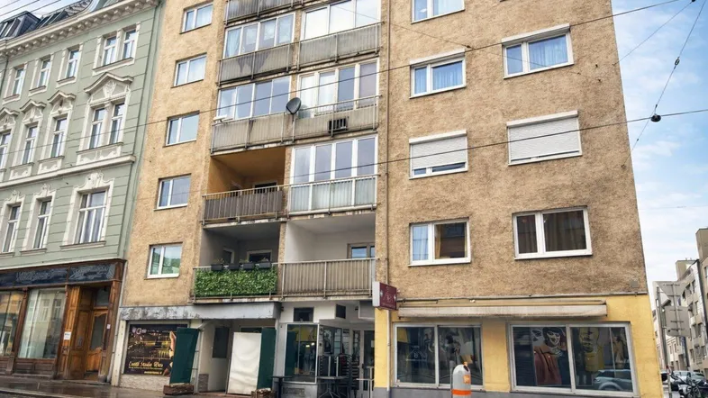 Mehrstöckiges Wohngebäude mit mehreren Balkonen und Geschäften im Erdgeschoss an einer Stadtstraße.