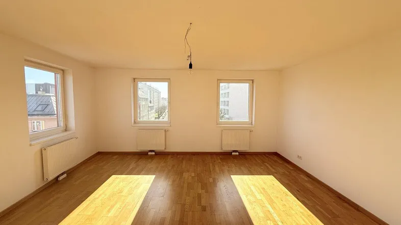 Helles, leeres Zimmer mit Parkettboden und mehreren Fenstern, das viel Sonnenlicht hereinlässt.
