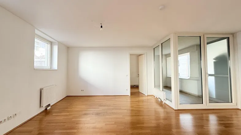 Heller Wohnbereich mit Parkettboden, weißen Wänden und Zugang zu einem verglasten Balkon.