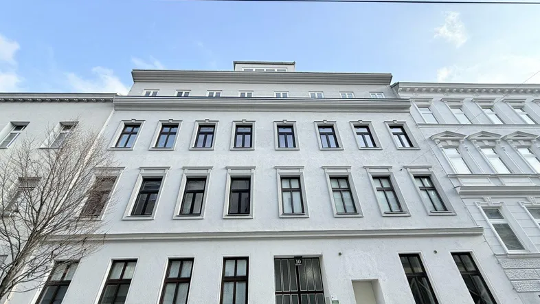 Die weiße Fassade des Wohngebäudes mit vielen Fenstern und dem Haupteingang.