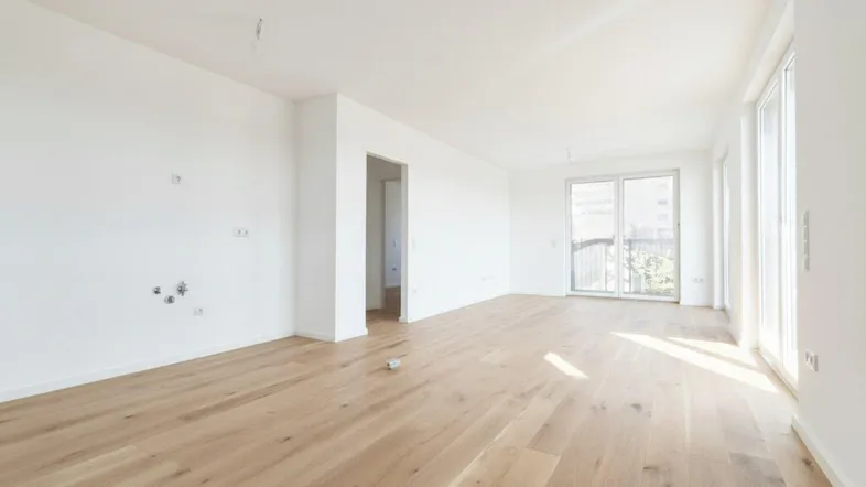 Helles, leeres Wohnzimmer mit neuem Parkettboden und Zugang zu einem Balkon.