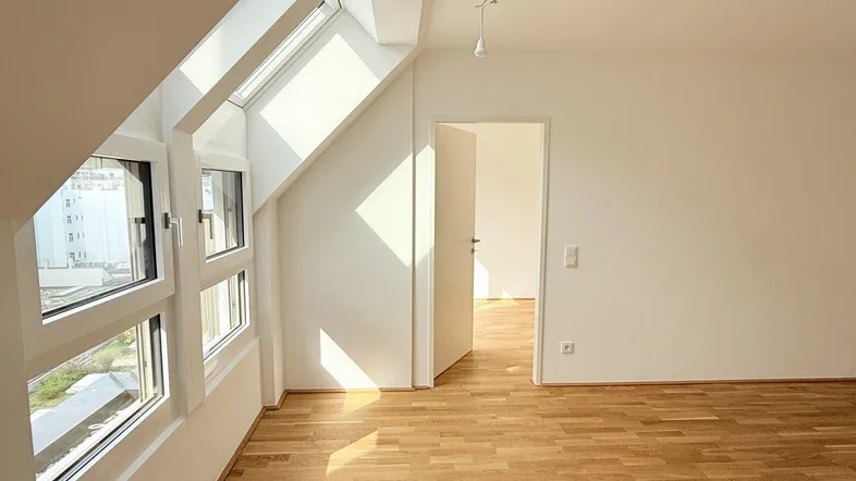 Helles Zimmer im Dachgeschoss mit schrägen Wänden, großen Fenstern und Parkettboden.