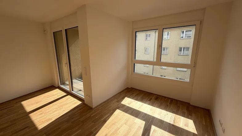 Helles Zimmer mit Parkettboden und Zugang zu einem Balkon, durchflutet von Sonnenlicht.