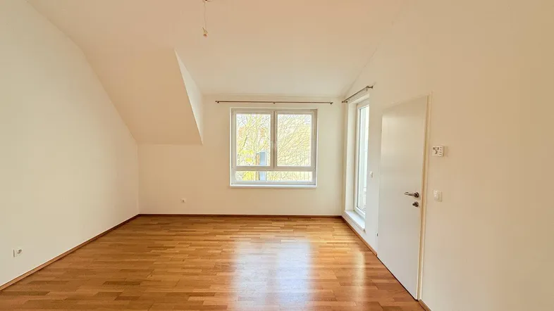 Helles Zimmer mit Parkettboden, weißen Wänden und einem Fenster, das viel Tageslicht hereinlässt.
