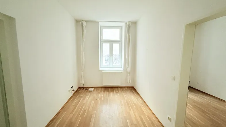 Heller, leerer Raum mit Holzparkettboden und einem Fenster, das Tageslicht hereinlässt.
