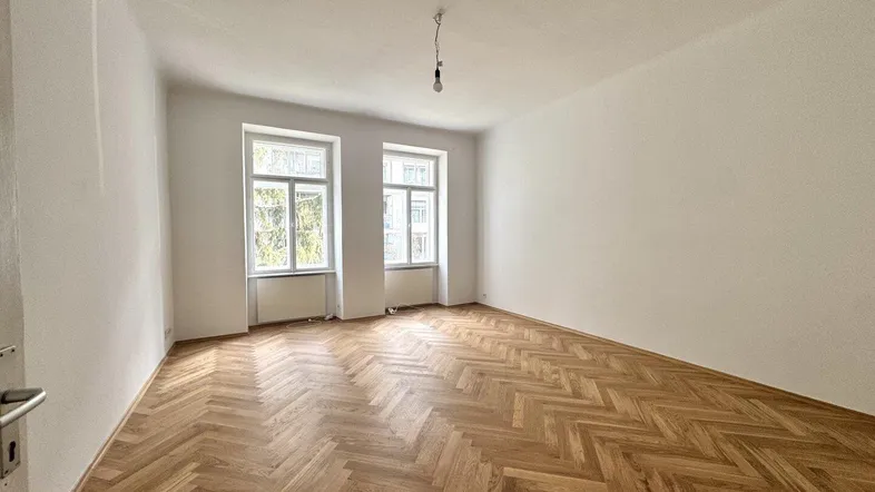 Helles Zimmer mit Fischgrätparkettboden und zwei großen Fenstern, ideal als Wohn- oder Schlafzimmer.