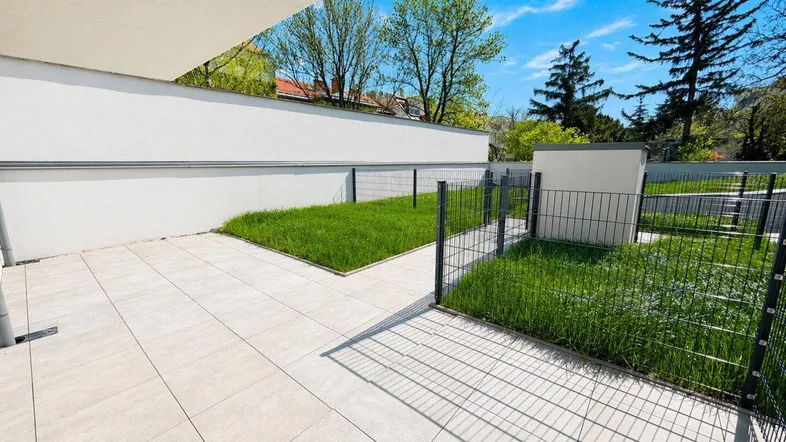 Geflieste Terrasse mit kleinen Rasenflächen und Zaun, umgeben von Grün und Bäumen.