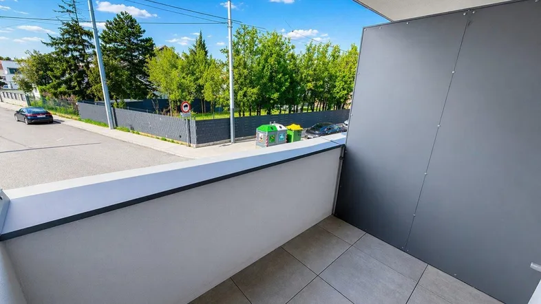 Heller Balkon mit Fliesenboden und Blick auf die Straße sowie umliegende Bäume.