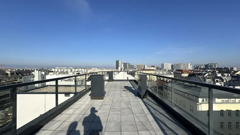 Geräumige Dachterrasse mit Fliesenboden und weitem Blick über die Stadt unter blauem Himmel.
