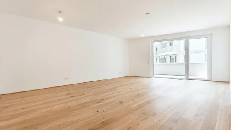 Helles, leeres Zimmer mit Holzboden und Zugang zu einem Balkon, ideal als Wohnbereich.