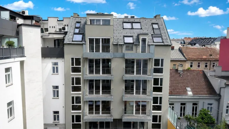 Moderne Wohnhausfassade mit vielen Fenstern, Balkonen und begrünten Dachflächen unter blauem Himmel.