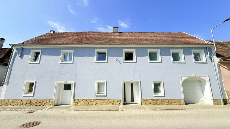 Hellblau gestrichene Fassade eines Mehrfamilienhauses mit braunem Ziegeldach und Steinsockel.