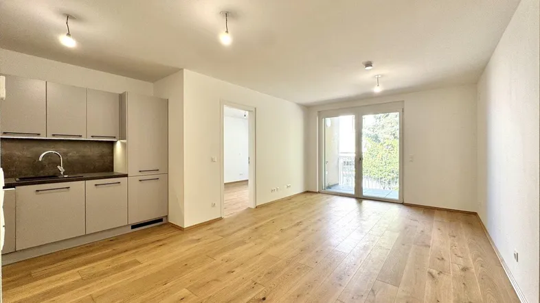 Heller offener Wohnbereich mit integrierter Küchenzeile, Holzboden und Zugang zum Balkon.