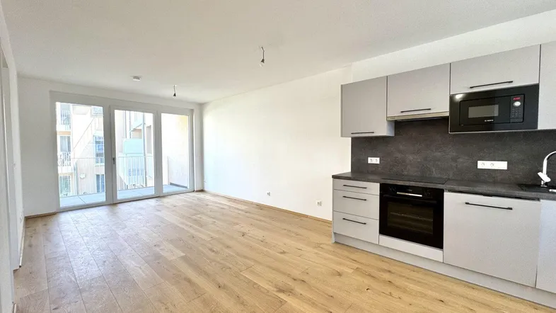 Heller offener Wohnbereich mit integrierter Küche, Holzboden und Zugang zu einem Balkon.