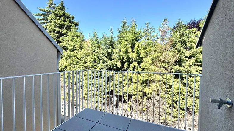 Heller Balkon mit Fliesenboden und Blick ins Grüne, umgeben von Bäumen.