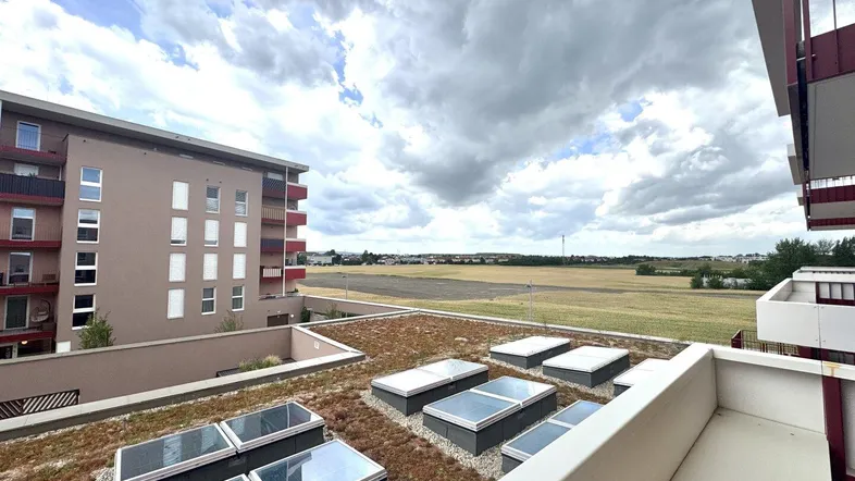 Blick von einem Balkon auf ein begrüntes Dach und ein weites Feld unter bewölktem Himmel.