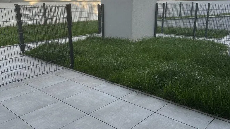 Gedeckte Terrasse mit Fliesenboden und angrenzender kleiner Rasenfläche, umzäunt von einem Metallzaun.