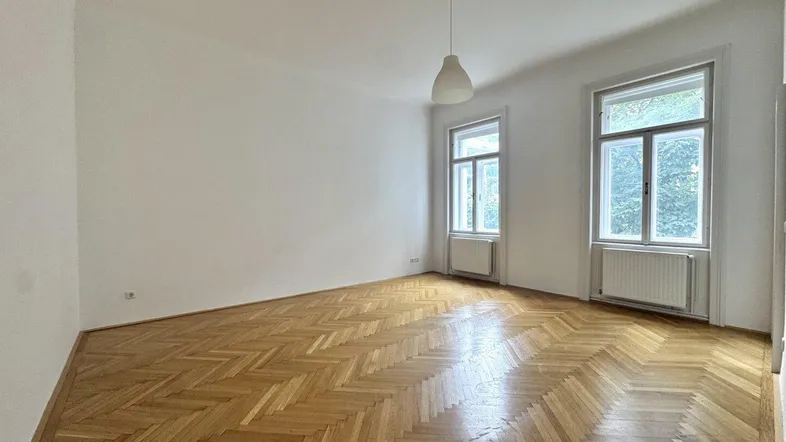 Geräumiges Wohnzimmer mit klassischem Fischgrätparkett und viel Tageslicht durch zwei Fenster.