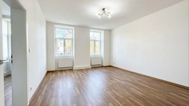 Helles, leeres Zimmer mit Holzboden und zwei großen Fenstern, bereit zur individuellen Gestaltung.
