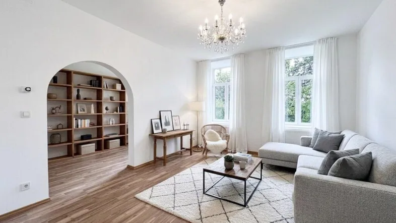 Helles Wohnzimmer mit Parkettboden, dekorativem Kronleuchter und einem offenen Durchgang zu einem weiteren Raum.