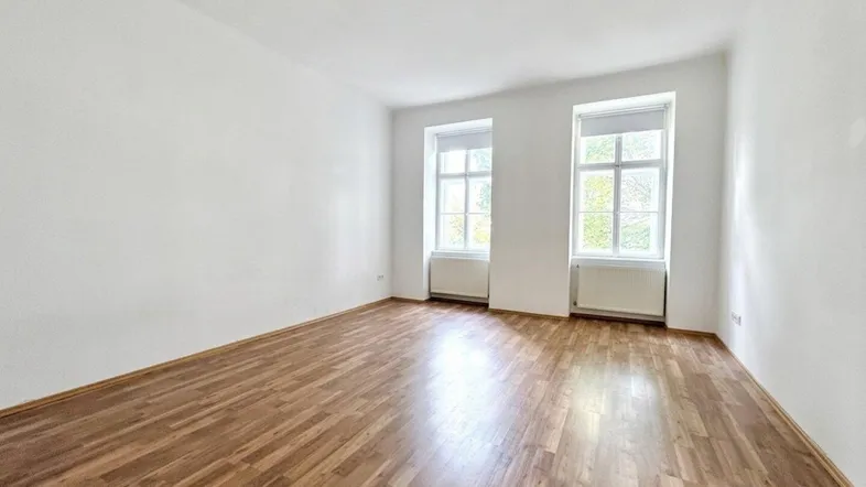 Heller, leerer Raum mit Holzboden und zwei großen Fenstern, ideal als Wohn- oder Schlafzimmer.