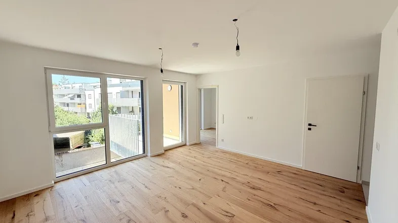 Helles, leeres Zimmer mit modernem Parkettboden und großen Fenstern, die Zugang zu einem Balkon bieten.