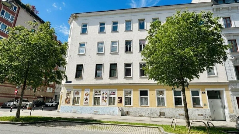 Ansicht eines Mehrfamilienhauses mit Geschäften im Erdgeschoss und Bäumen an der Straße.