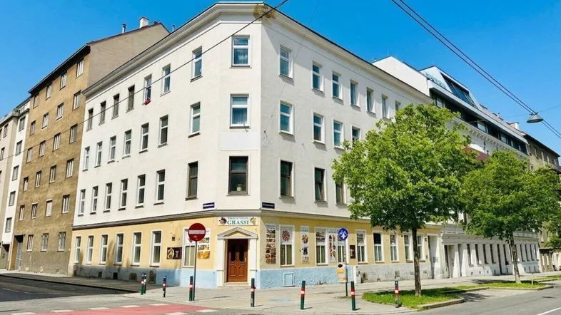 Helles Eckgebäude in städtischer Umgebung mit Geschäften im Erdgeschoss und gepflegter Fassade.