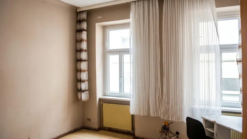 Helles Zimmer mit beigen Wänden, Holzfußboden und einem Fenster mit Vorhängen und Heizkörper.
