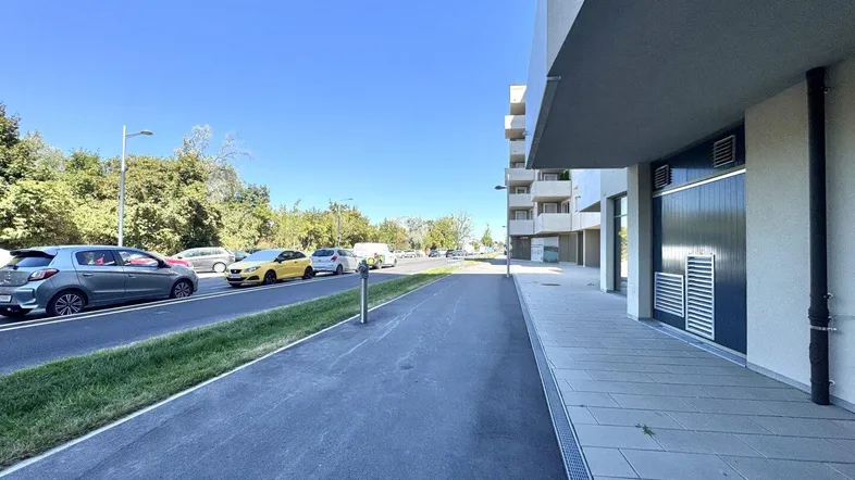 Moderne Gebäudefassade mit Bürgersteig, Parkplätzen und Bäumen im Hintergrund unter blauem Himmel.