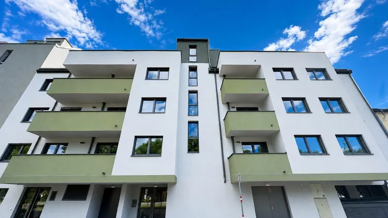 Moderne Wohnhausfassade mit weißen Wänden und grünen Balkonen unter blauem Himmel.